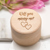 Wooden box with 'Will you marry me?' engraving on a white surface with pink flowers in the background