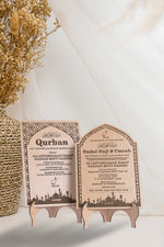 Two Qurban invitations on wooden stands with dried flowers and a vase in the background.